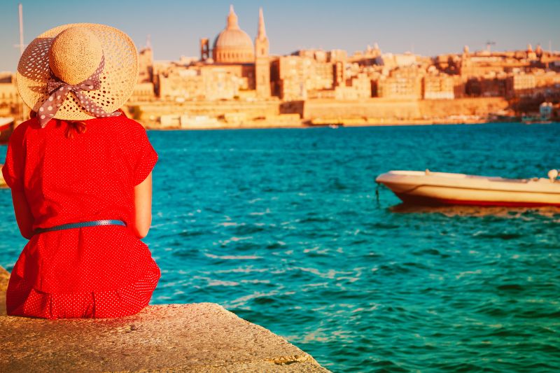 A woman in a flowing red dress sits by the sea in Malta, gazing at the stunning view of Valletta's historic skyline. The golden limestone buildings contrast beautifully with the deep blue waters, creating a picturesque and serene scene.